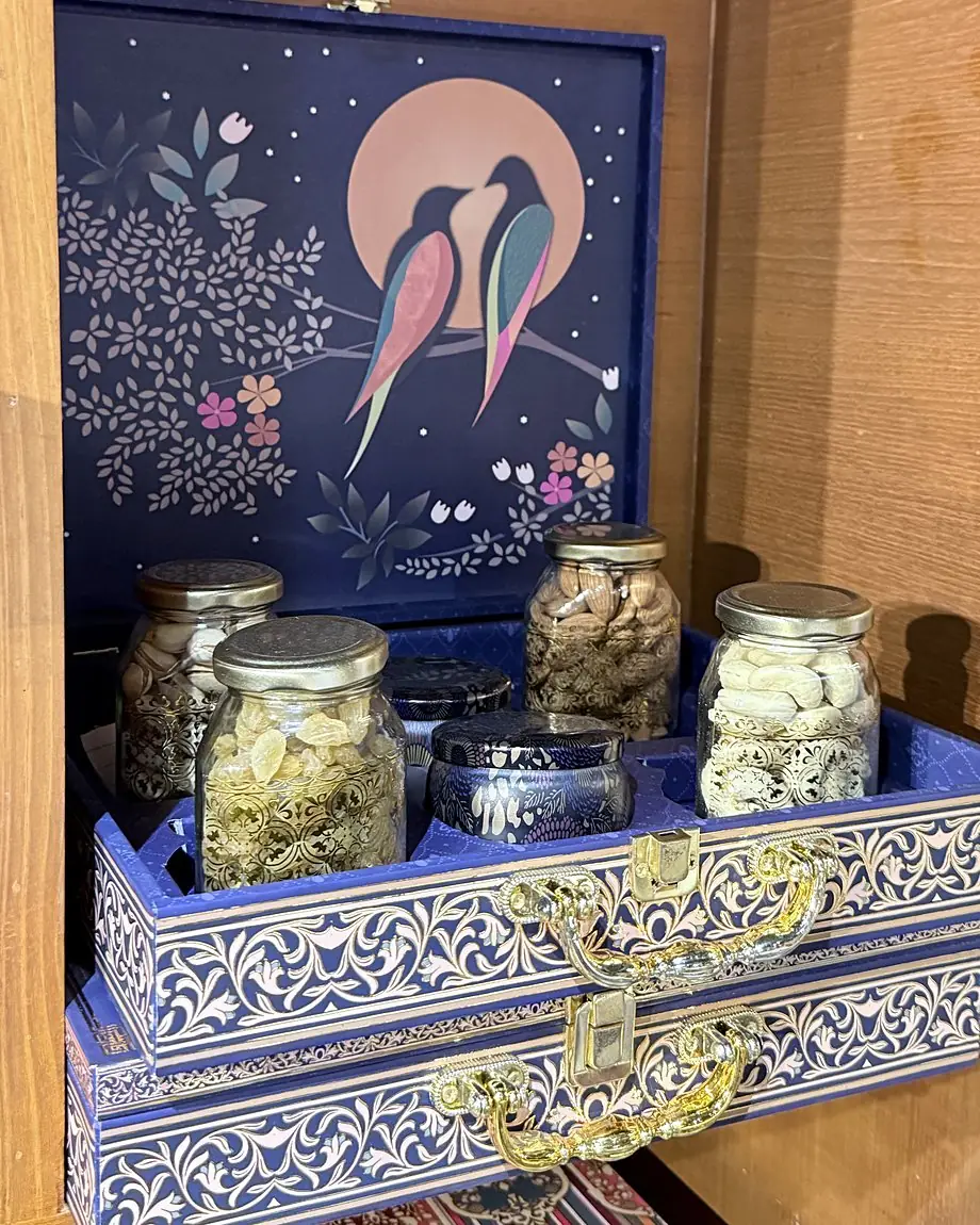 An open navy gift box with two birds against a full moon painted on the lid, holding glass jars of cashews, almonds and other dry fruits