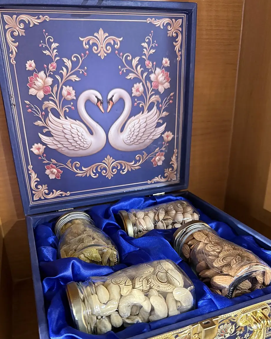 An open blue gift box with two white swans on the lid and four glass jars of cashews, almonds, pistachios and raisins on blue satin