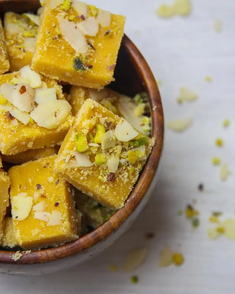 Squares of barfi topped with almond and pistachio in a wooden bowl