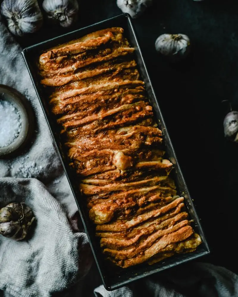 A scored garlic bread loaf in a baking tin, herbs through the cuts