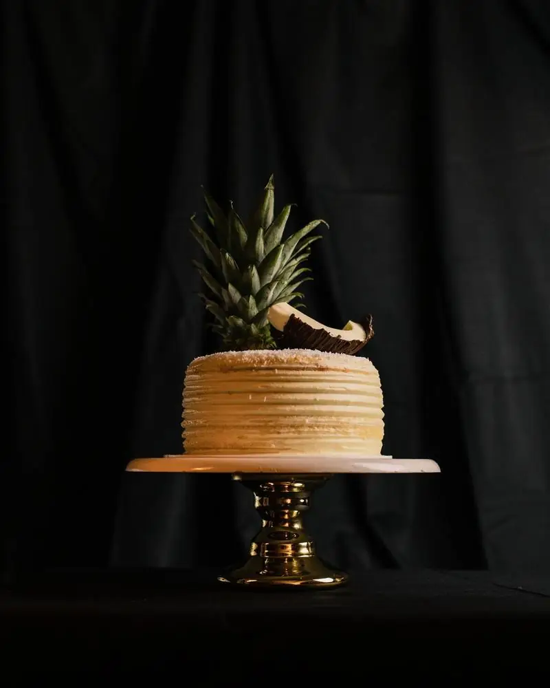 A pineapple cream cake on a raised stand, topped with a pineapple crown